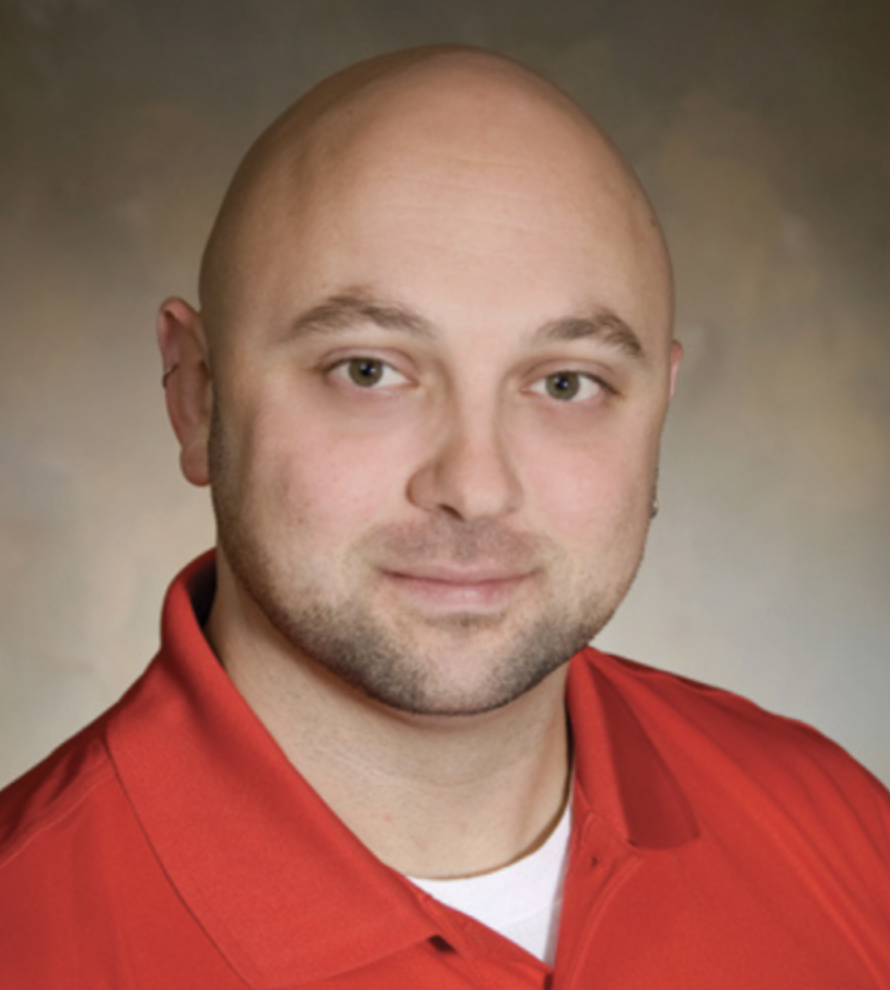 Professional headshot of a bald man with a trimmed beard, wearing a red collared shirt, looking confidently at the camera. The background is softly blurred in neutral tones, conveying a friendly and approachable healthcare professional.