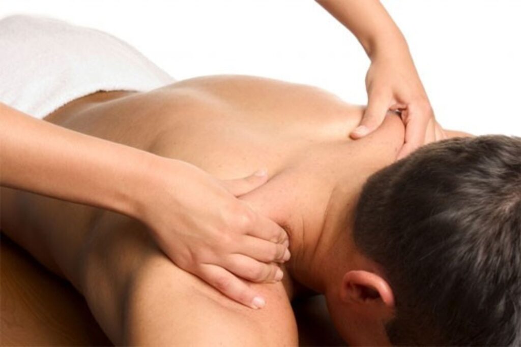 A practitioner applies firm pressure to a man’s upper back and shoulder while he lies face down on a treatment table. The hands on technique focuses on the neck and shoulder area, illustrating massage relieving muscle tension and improving mobility.
