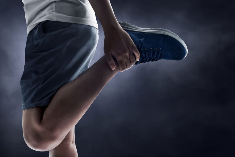 Person in athletic shorts and blue running shoes stretches their leg by pulling one foot up behind them before exercise. The image represents mobility and joint care often associated with osteopathy support and physical wellness.